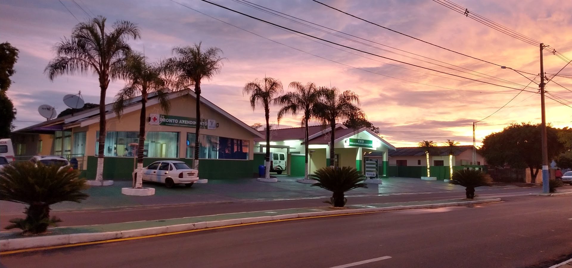Vista da Santa Casa de Quintana a partir da avenida, com céu alaranjado do pôr do sol e ambulância estacionada na entrada.