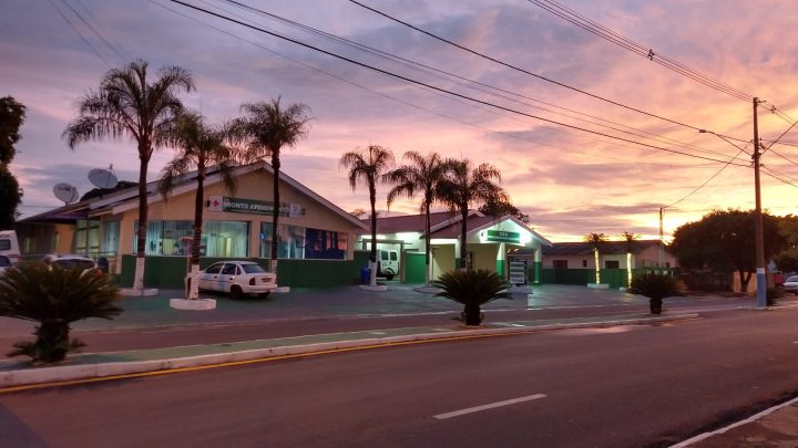 Vista da Santa Casa de Quintana a partir da avenida, com céu alaranjado do pôr do sol e ambulância estacionada na entrada.